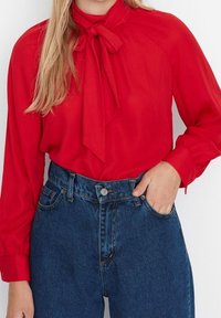 Red blouse with a bow tie at the neckline, long sleeves, and smooth fabric, paired with high-waisted blue denim jeans, featuring front pockets.