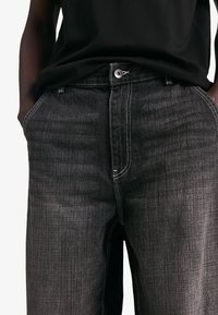 Pantaloni negri din denim cu croială dreaptă, având cusături albe, închidere cu nasture și buzunare laterale. Materialul are o textură ușoară.