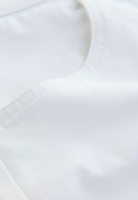 Tessuto bianco con una texture liscia, dotato di scollatura curva e dettagli di cucitura precisi. Il materiale appare leggero e morbido.