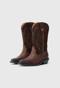 Pruun nahast ja sametist cowboy saapad, millel on dekoratiivne valge tikand, terav nina ja madal, virnastatud konts. Omab tõmbeotsi, mis võimaldab neid kergesti jalga panna.