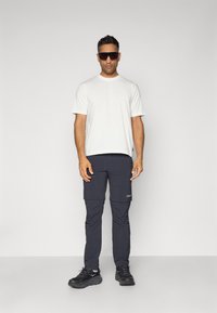 White short-sleeve shirt, dark cargo pants with pockets, and black shoes. Individual is wearing oversized black sunglasses.