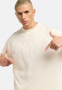 Beige hettegenser med opphøyd broderi av "Colucci la Famiglia" på fronten, med raglanermer og rund halsdesign.