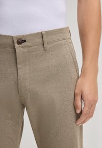 Pantaloni beige con texture, chiusura a bottone, frontale piatto e tasche laterali. Il tessuto presenta un motivo intrecciato, evidenziando un design informale.