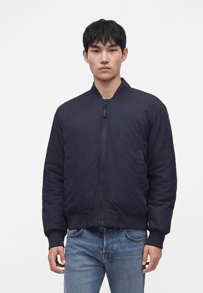 Homme portant une veste bomber noire zippée et un jean bleu, debout devant un fond blanc uni.