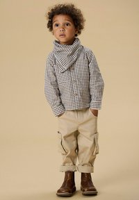 Hnědá kostkovaná košile na knoflíky, béžové kalhoty cargo s ohrnutými nohavicemi, hnědé kožené boty, doplněné odpovídajícím šátkem. Jednoduchý a praktický design.