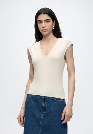 Joven mujer con cabello largo y oscuro lleva top sin mangas beige con escote en V y falda de denim azul sobre un fondo liso y claro.