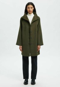 Olijfgroene wollen jas met een brede kraag, knoopsluiting aan de voorkant en oversized mouwen, gecombineerd met donkere jeans en zwarte schoenen.