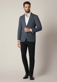 Szary, teksturowany żakiet z wyciętymi klapami, biała koszula pod spodem, czarne spodnie i czarne buty eleganckie, stojący na neutralnym tle.