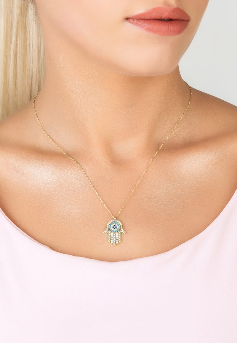 Guldkæde med en hamsa vedhæng, prydet med blå og klare rhinestones, vist mod en lys pink top.