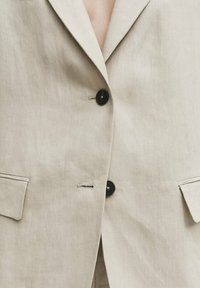 Veste en lin beige clair à coupe simple boutonnage, avec deux poches avant et des boutons noirs pour le contraste. Texture lisse.