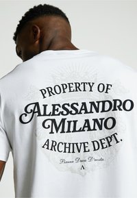 Valkoinen puuvillapaita, jossa on mustaa tekstiä selkäpuolella: "ALESSANDRO MILANO -ARKISTOSIIRTO." koristeellisin elementein.