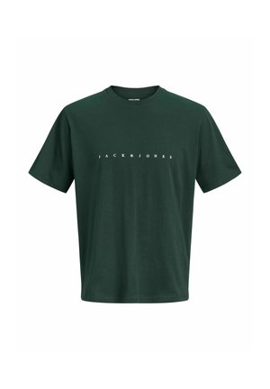 Jongen staat frontaal, draagt een geruit overhemd over een donkergroen T-shirt, een donkere spijkerbroek en beige schoenen op een effen achtergrond.