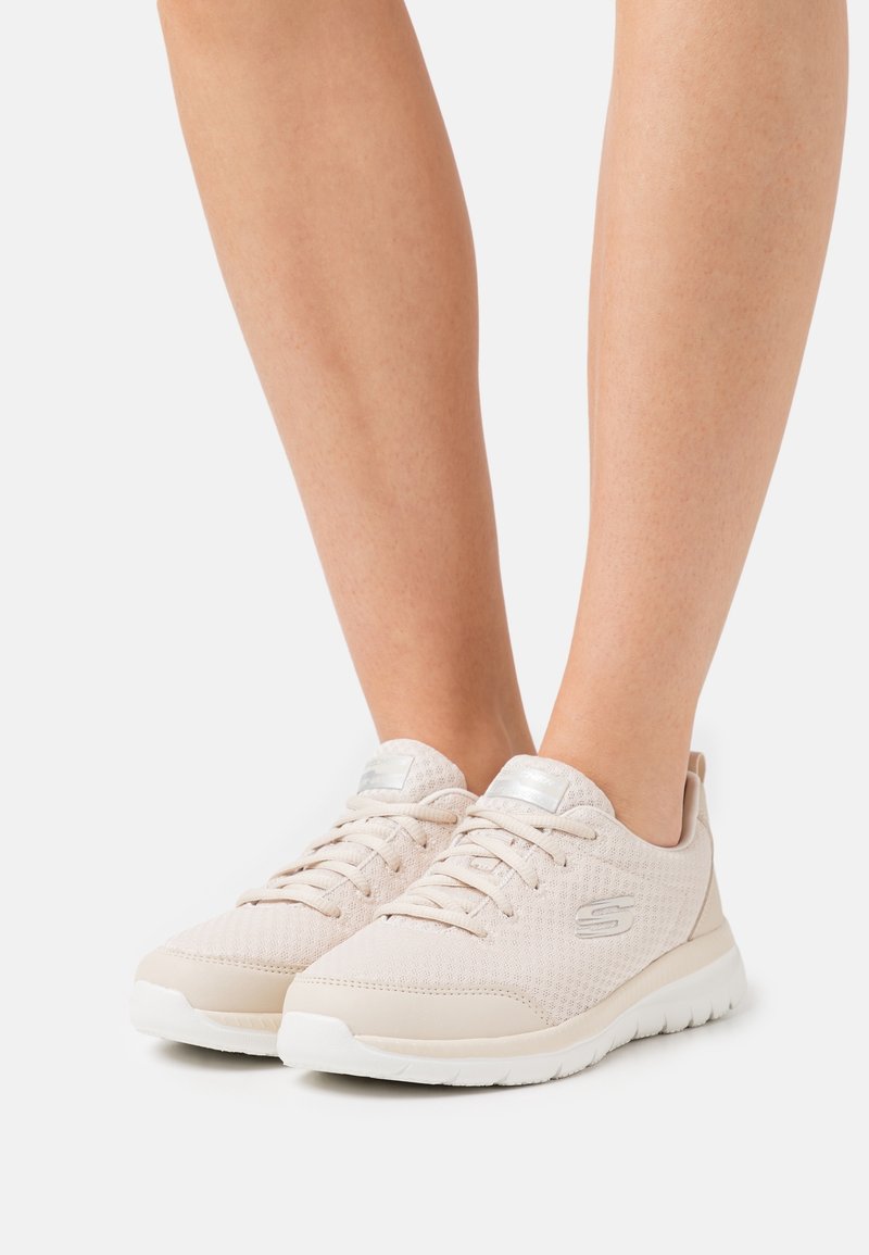 Jambes portant des baskets en mesh beige clair Skechers avec semelles blanches, debout sur une surface blanche devant un fond uni.