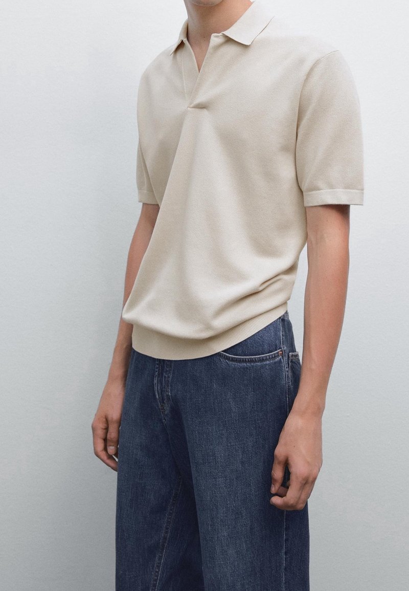 Personne portant un polo en maille beige à manches courtes, associé à un jean bleu foncé ample, sur un fond clair uni.