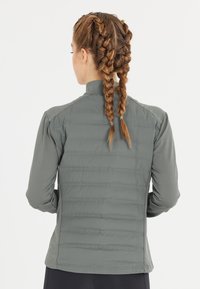 Grå jakke med en tætsiddende design, der har en quiltet tekstur på ryggen og glatte ærmer. Høj krave og lange ærmer.