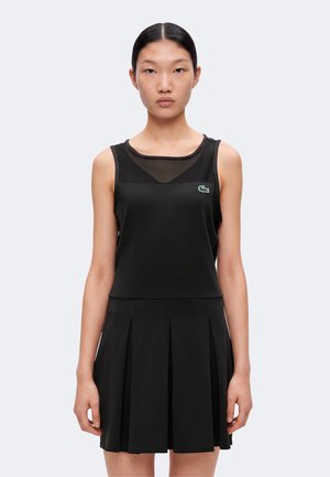 Joven mujer con el pelo negro y liso lleva un vestido negro sin mangas con falda plisada y un pequeño logo verde de cocodrilo en el pecho.