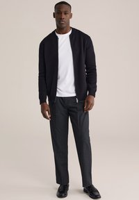 Juoda bombonešio striukė su užtrauktuku dėvima ant baltos marškinėlių, derinama su pilkomis kelnėmis ir juodais batais. Paprastas dizainas, lygi tekstūra.