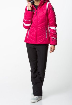 Růžová prošívaná bunda s bílými pruhy, se zapínacími kapsami a kapucí lemovanou kožešinou, spárovaná s černými kalhotami a světlými botami.