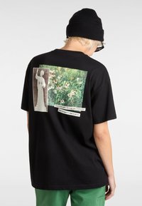 Sort bomulds t-shirt med et stort grafisk motiv på ryggen, der viser to billeder: en statue og en blomsterscene, ledsaget af tekstoverskrifter.