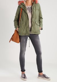Olivgrüne Parka mit rosa Pelzkapuze, beige gestrickter Pullover, graue Skinny-Jeans und metallische Slipper. Braune Umhängetasche.