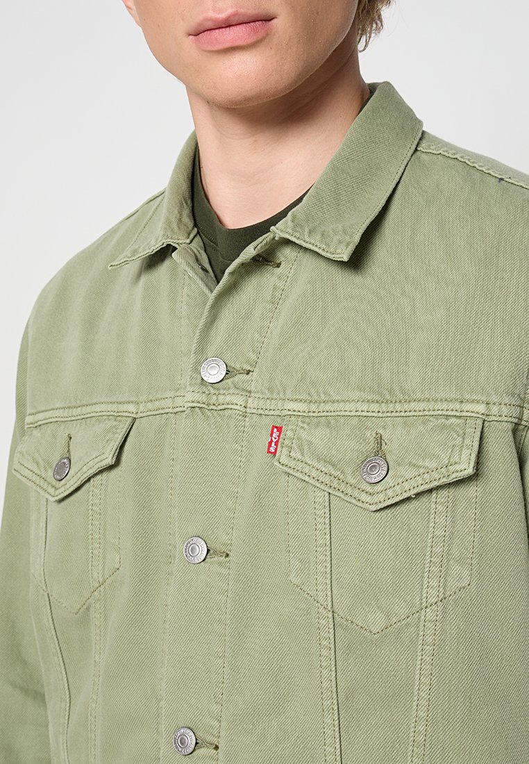 Giacca di jeans verde chiaro con collo a camicia, bottoni a pressione, tasche sul petto e una piccola etichetta rossa sul taschino sinistro.
