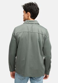 Grøn langærmet skjorte med krave, tekstureret stof og syede skulderaccenter. Båret over lyseblå jeans, vist fra ryggen.
