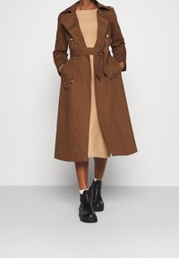 Cappotto trench marrone con vita cinturata, tasche laterali e colletto a revers, indossato sopra un vestito beige e abbinato a stivali neri chunky alla caviglia.