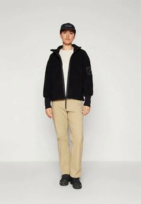 Schwarze Fleece-Jacke mit Reißverschluss und Stehkragen, kombiniert mit einem hellbeigen Hemd und beigen geraden Hosen, abgerundet mit schwarzen Schuhen.