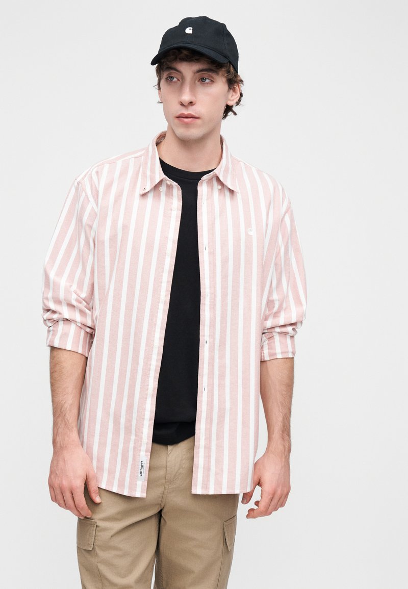 Chemise à rayures boutonnée en rose clair et blanc, manches longues retroussées, associée à un t-shirt noir et une casquette, ainsi qu'à un pantalon cargo kaki.
