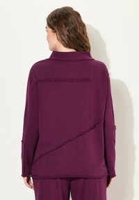 Volná, oversize fialová košile s límečkem, která má asymetrický design, třepené okraje a texturovanou látku.