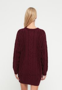 Mahonie kabelgebreide jurk met lange mouwen, een ronde halslijn en een geribde zoom. De textuur is grof met ingewikkelde patronen.