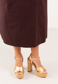 Sandalias de plataforma doradas con tiras cruzadas y hebilla en el tobillo. Base tejida texturizada y tacón bloque. Color neutro que acentúa el diseño.