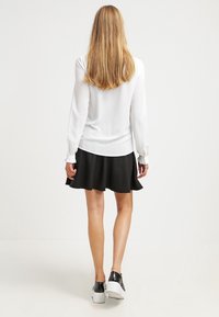 Kvinde med langt blondt hår iført en hvid bluse med lange ærmer, sort nederdel og sorte platform shoes, står med ryggen til foran en ensfarvet baggrund.