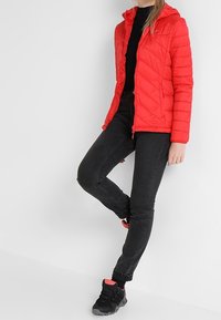 Červená prošívaná bunda s kapucí, zapínáním na zip a prošitým vzorem; nošena přes černý top s černými úzkými džíny a černými teniskami s růžovými akcenty.