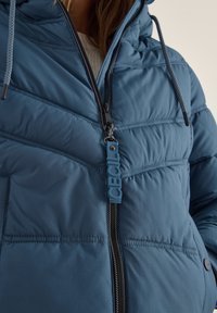 Giubbotto imbottito blu con chiusura a zip, superficie testurizzata, cappuccio e dettaglio logo sul tirante della zip. Presenta sezioni cucite per l'isolamento.