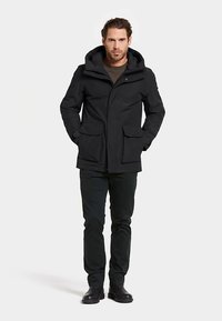 Musta vedenpitävä takki hupulla, jossa on edessä vetoketju, kaksi sivutaskua ja rennosti istuva leikkaus, yhdistetty tummien housujen ja saappaiden kanssa.