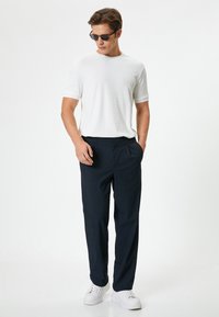 Witte gestructureerde kortemouwen shirt gecombineerd met marineblauwe broek en witte sneakers. Model draagt een donkere zonnebril, met de handen in de zakken.