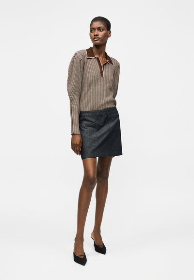 Ψηλή γυναίκα που φοράει μακρυμάνικη μπλούζα με καφέ ρίγες και γιακά, μαύρη τζιν φούστα και μαύρες μυτερές γόβες, στέκεται με χαλαρά χέρια.