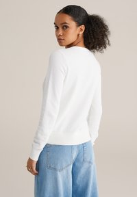 Pulover alb texturat cu gât rotund și mâneci cu tivuri striate, asortat cu blugi largi de culoare albastru deschis. Design simplu și minimalist.