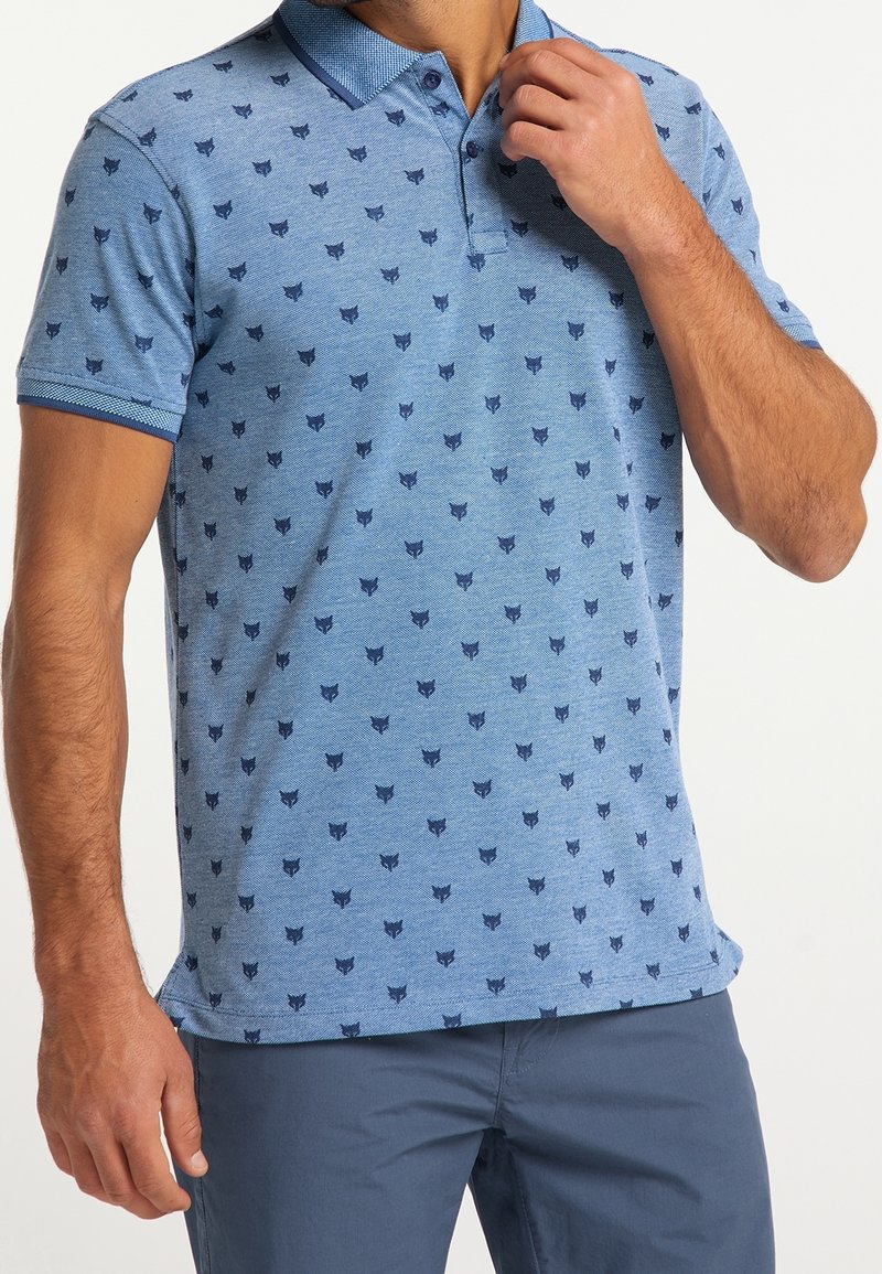 Homme portant un polo bleu clair avec des motifs de têtes d’animaux bleu foncé et un pantalon bleu assorti, ajustant le col de la chemise.