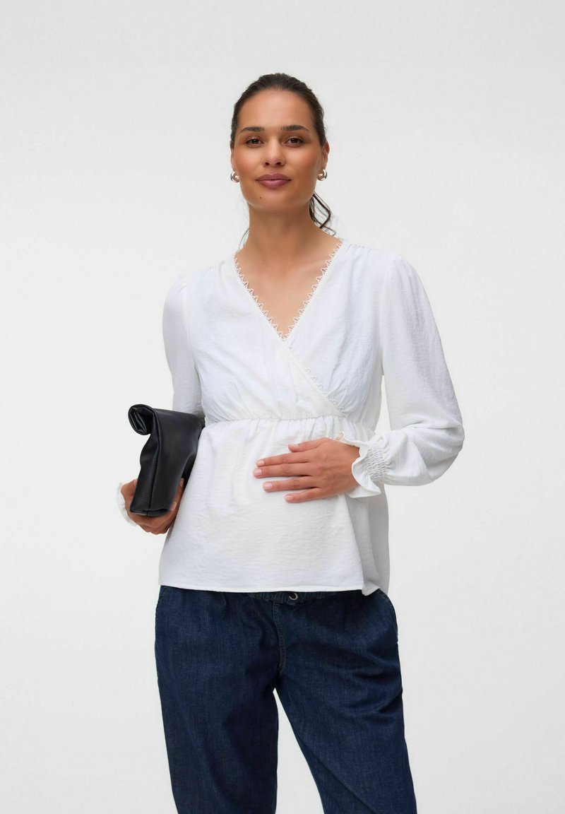 Femme portant un chemisier blanc à manches longues et un jean foncé, tenant une pochette noire dans une main et touchant son abdomen de l'autre.