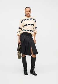 Γυναίκα που φοράει μπεζ πουλόβερ με μαύρες καρδιές, μαύρη πλισέ φούστα με πλαϊνή σκίσιμο, μαύρες μπότες μέχρι το γόνατο και κρατάει μια τσάντα ελιάς.