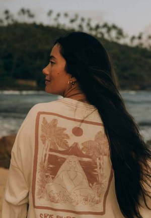 Kobieta z długimi ciemnymi włosami, ubrana w kremową koszulę z brązowym nadrukiem przedstawiającym scenę natury, stojąca w pobliżu plaży, w tle rozmyte drzewa.