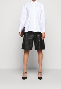 Camicia bianca a maniche lunghe, abbinata a pantaloni bermuda in pelle nera. Ulteriori dettagli includono una piccola tasca sul petto e eleganti tacchi neri.
