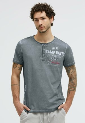 Junger Mann mit lockigem Haar, der ein ausgewaschenes graues Camp David T-Shirt trägt, mit Tattoos auf beiden Armen, Hände in den Taschen einer hellen Hose.