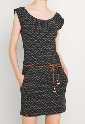 Nainen, jolla on musta mekko harmaalla siksak-kuviolla, lyhyet hihat, oranssi letitetty vyö helmillä ja taskut molemmilla puolilla.