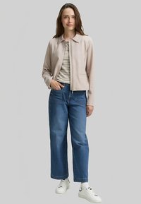 Hellrosa Reißverschlussjacke mit Kragen, darunter ein gestreiftes Hemd, kombiniert mit locker sitzenden blauen Jeans und weißen Turnschuhen. Einfaches Design.