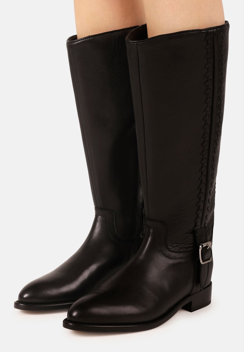 Bottes hautes en cuir noir avec une boucle latérale et des détails cousus, portées sur des jambes nues, sur un fond clair uni.