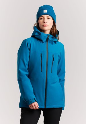 Vrouw met een blauwe gewatteerde jas met capuchon en met ritssluitingen voorziene zakken en een bijpassende blauwe muts, staand tegen een effen lichte achtergrond.