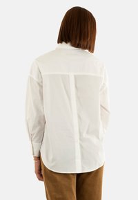 Chemise blanche à manches longues avec une coupe droite, dotée d'une couture de dos et de poignets à boutons, associée à un pantalon en velours côtelé marron.